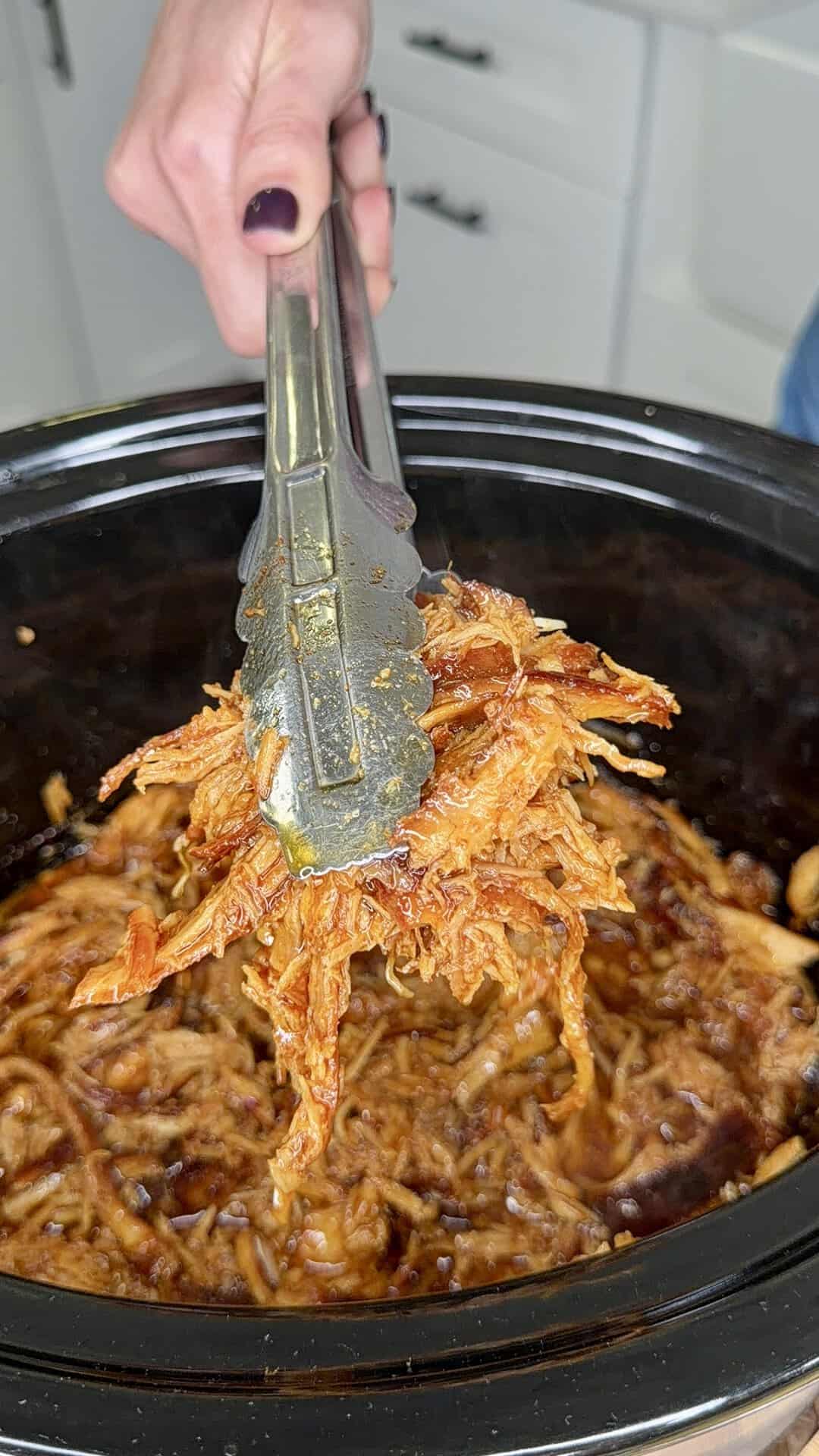 A hand holding metal tongs lifts a portion of saucy, shredded pulled pork from a black slow cooker filled with more pulled pork. Kitchen cabinets are visible in the background.