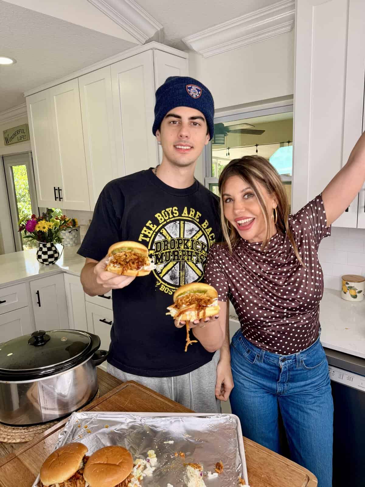 A young man in a black beanie and a smiling woman in a polka dot shirt stand in a bright kitchen, each holding a pulled pork sandwich. Sandwiches and a slow cooker are on the counter in front of them.