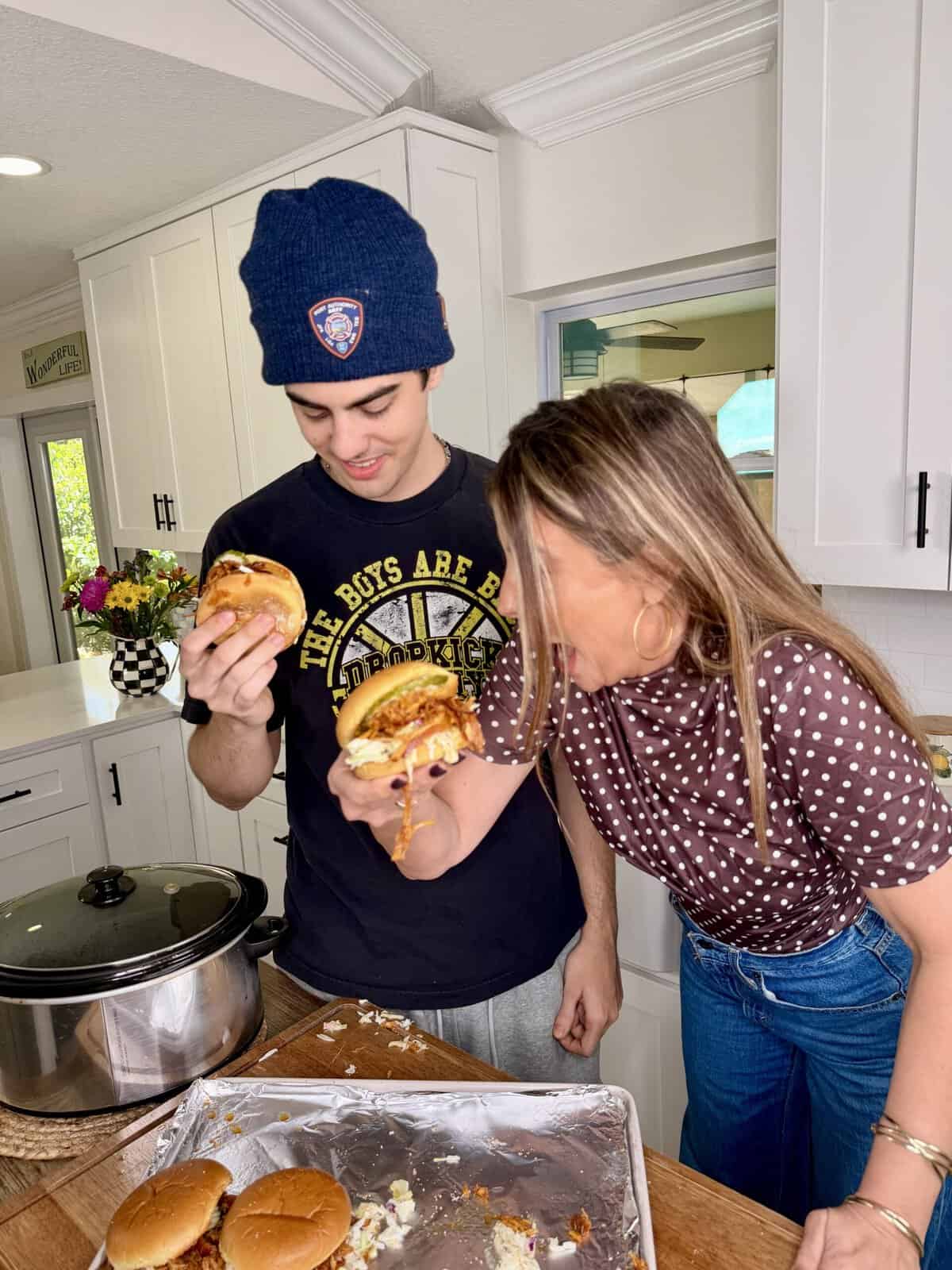 Two people in a kitchen hold large, messy burgers and smile at each other. A tray with more burgers and a slow cooker are on the counter in front of them. The setting is bright and casual.