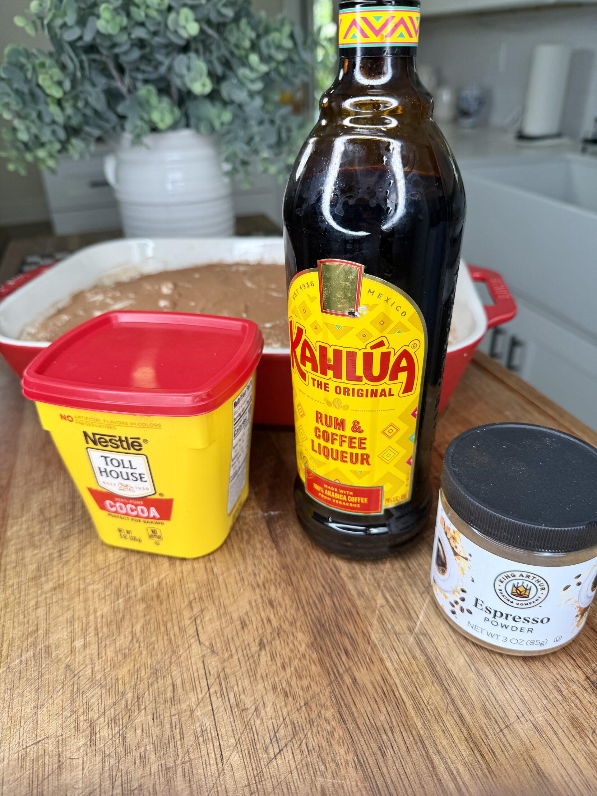 A container of Nestlé Toll House cocoa, a bottle of Kahlúa, and a jar of espresso powder are on a wooden surface, with a chocolate batter in a red dish and a plant in the background.