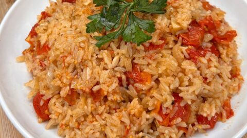 A white plate filled with tomato rice garnished with fresh parsley, served on a wooden surface. The rice is mixed with chopped tomatoes and appears fluffy and vibrant.