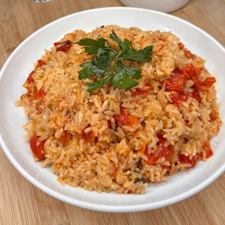 A white plate filled with tomato rice garnished with fresh parsley, served on a wooden surface. The rice is mixed with chopped tomatoes and appears fluffy and vibrant.