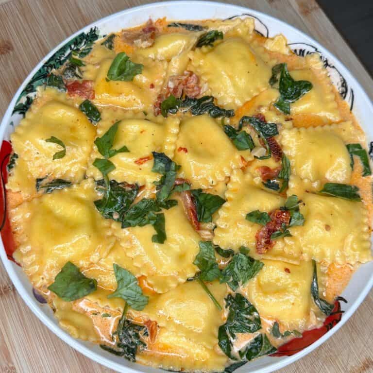 A bowl filled with cheese ravioli in a creamy orange sauce, garnished with fresh spinach, sundried tomatoes, and chopped parsley.