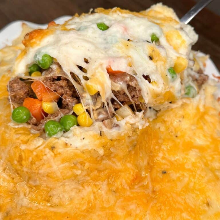 A close-up of a serving of shepherd’s pie, showing cheesy mashed potato topping, ground meat, peas, corn, and carrots inside. A spoon is lifting a portion, revealing the filling and melted cheese.