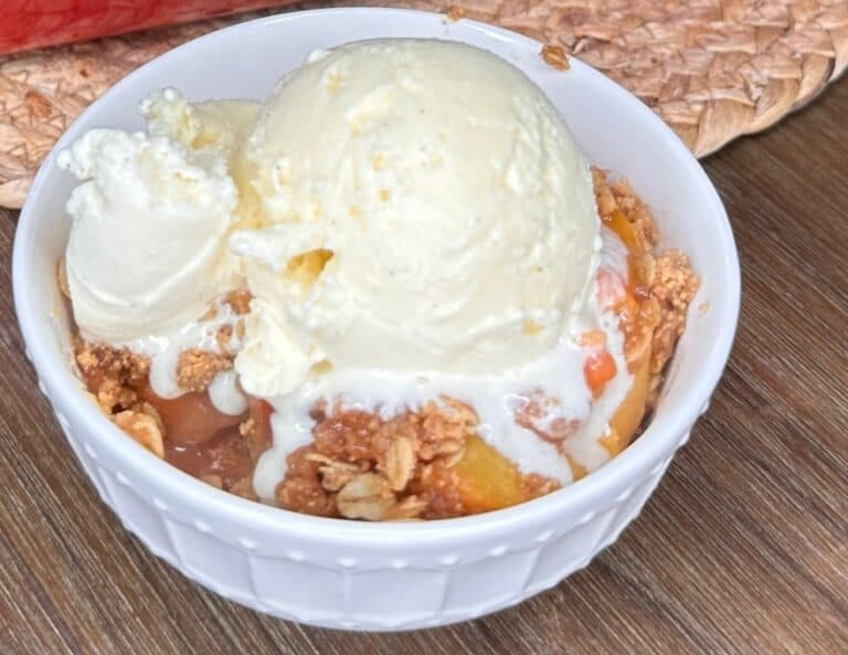 A white bowl filled with peach cobbler topped with a scoop of vanilla ice cream, starting to melt over the warm dessert, placed on a wooden surface.