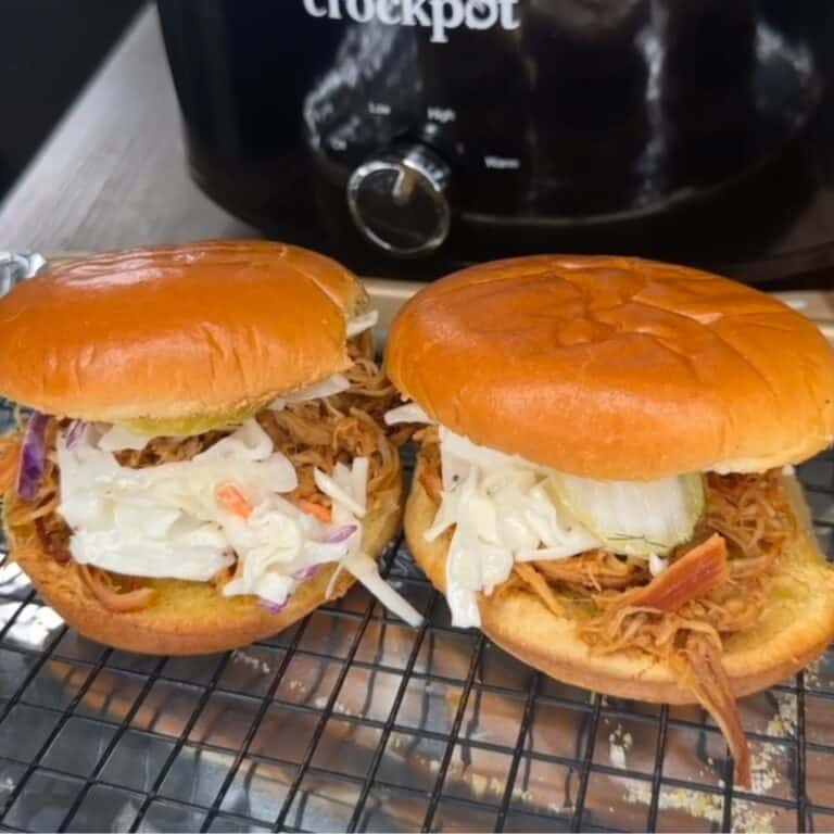 Two pulled pork sandwiches with coleslaw on brioche buns are placed on a cooling rack. A slow cooker labeled “crockpot” is in the background.