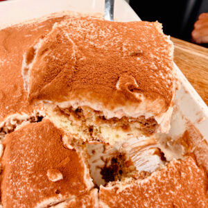 A close-up of a tray of tiramisu with a large square piece being lifted out, showing layers of cream and coffee-soaked cake, topped with cocoa powder.