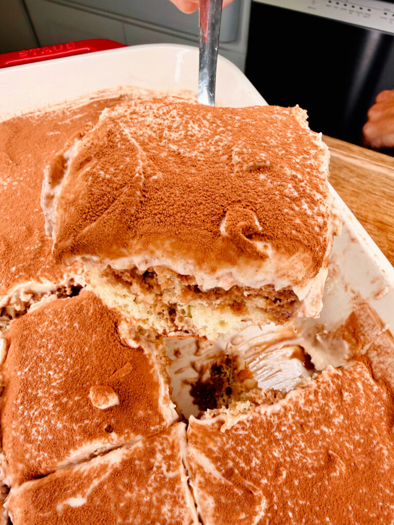 A close-up of a tray of tiramisu with a large square piece being lifted out, showing layers of cream and coffee-soaked cake, topped with cocoa powder.
