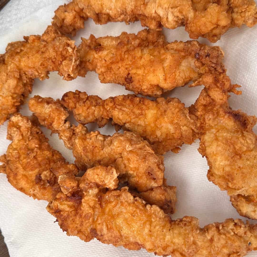Several crispy, golden brown fried chicken tenders rest on a white paper towel, showing their crunchy, textured coating.