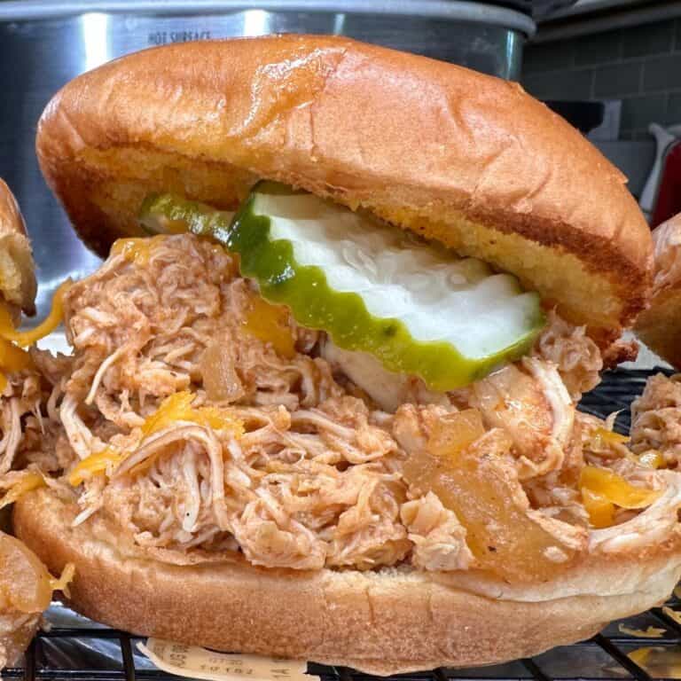 A close-up of a pulled chicken sandwich on a toasted bun, topped with pickles and melted cheese, with a slow cooker and kitchen tiles visible in the background.