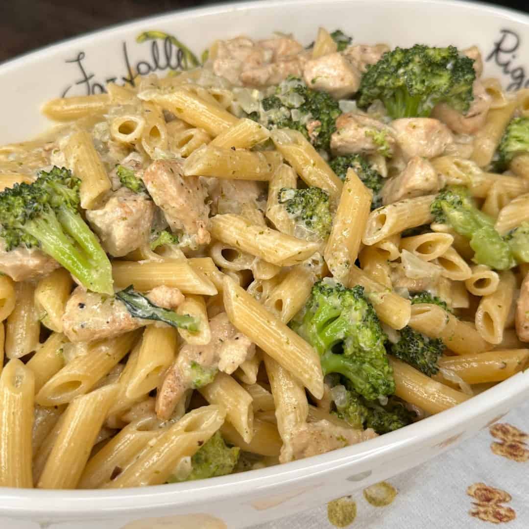 A close-up of creamy penne pasta mixed with broccoli florets and chunks of seasoned chicken, served in a white casserole dish.