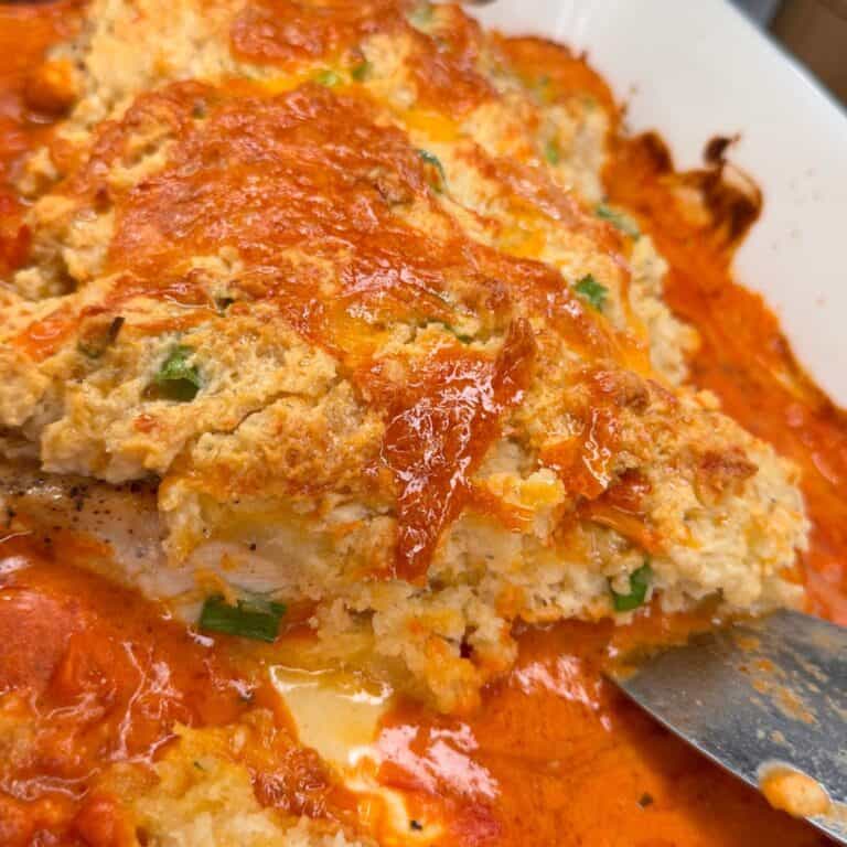 A close-up of a cheesy, baked casserole with a golden crust, visible green herbs, and a thick layer of melted red-orange sauce, with a serving spoon in the corner of the dish.