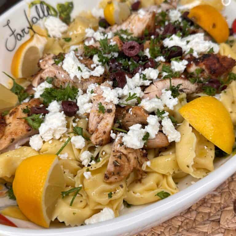 A plate of bow-tie pasta topped with grilled chicken, crumbled feta cheese, sliced black olives, fresh parsley, and lemon wedges, served on a white dish.