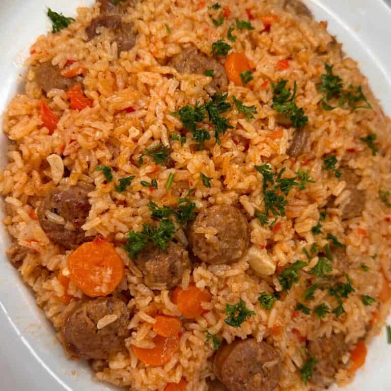 A close-up of a plate of jollof rice with slices of sausage, carrots, and garnished with fresh parsley. The rice is orange-red, indicating a tomato-based sauce, and vegetables and meat are mixed throughout.