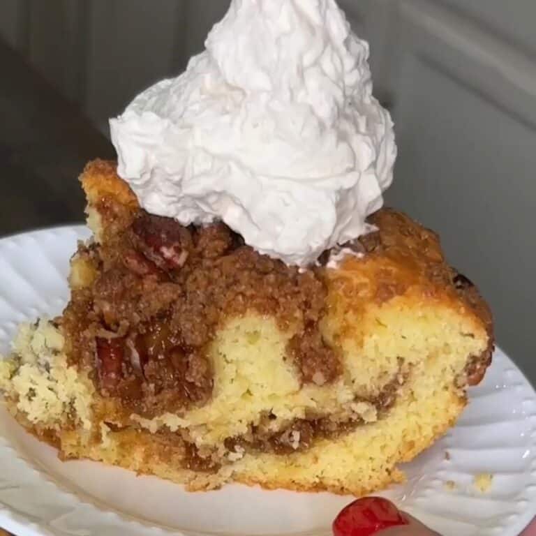 A slice of coffee cake with a crumbly cinnamon filling and pecans, topped with a large dollop of whipped cream, served on a white plate.
