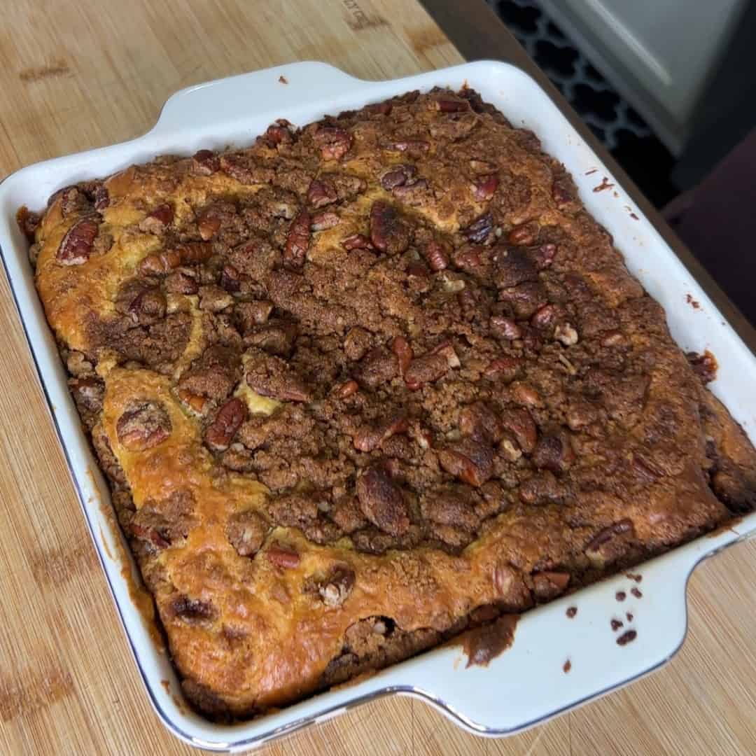 A freshly baked coffee cake topped with a layer of chopped nuts and crumbly brown sugar streusel, sitting in a white square baking dish on a wooden surface.