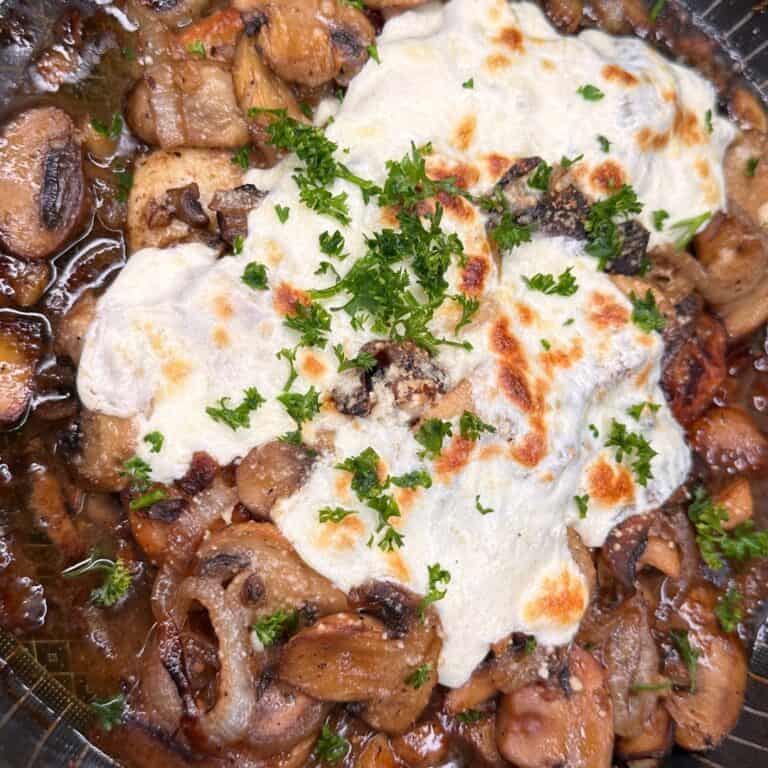 A close-up of a skillet filled with sautéed mushrooms and onions, topped with melted cheese and garnished with chopped parsley.