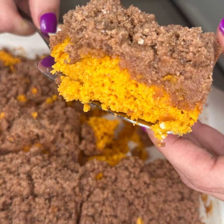 A hand with purple nail polish holds a slice of cake with a bright orange base and a crumbly brown topping, with more cake visible in the background.