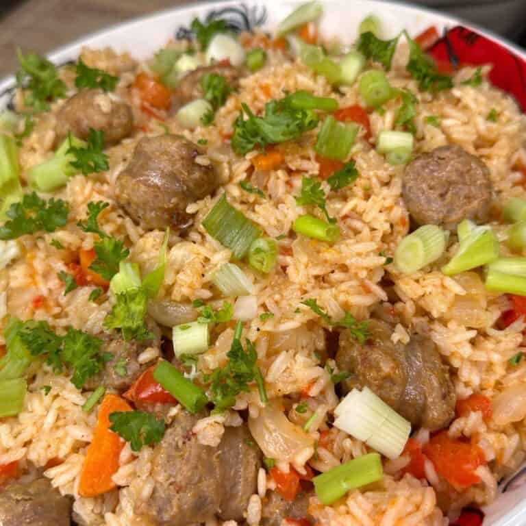 A close-up of a dish featuring seasoned rice mixed with chunks of sausage, diced red bell peppers, chopped green onions, and fresh parsley. The ingredients are vibrant and well-mixed in a patterned bowl.