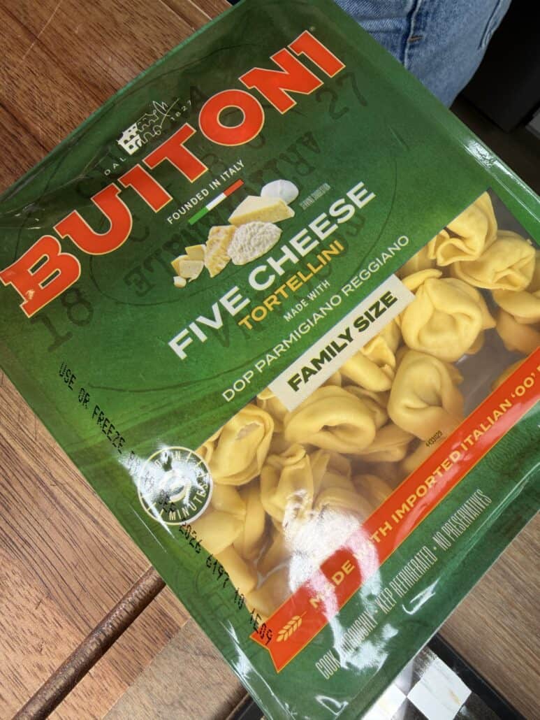 A package of Buitoni Five Cheese Tortellini, family size, is shown on a wooden surface. The green package features images of cheese and pasta, labeled DOP Parmigiano Reggiano..
