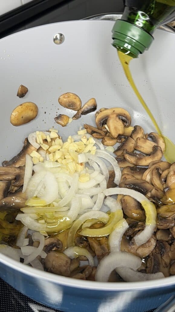 Sliced mushrooms, onions, and garlic are sautéing in a pot, with olive oil being poured in from a green bottle.