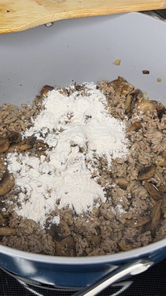 Ground meat and chopped mushrooms are cooking in a pan, with a mound of white flour sprinkled on top, ready to be stirred in. A wooden spoon rests on the pans edge.