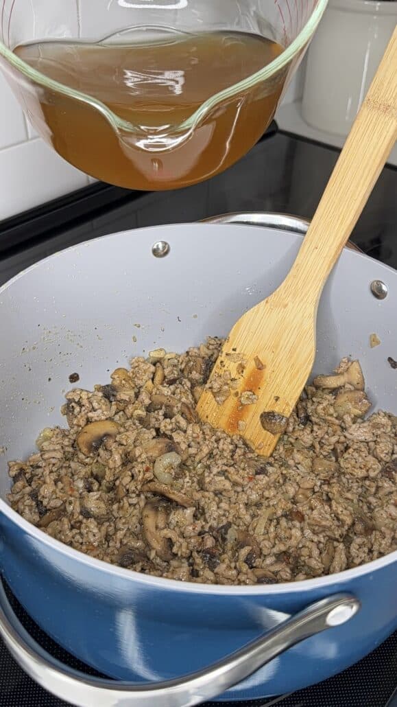 Ground meat and mushrooms cooking in a blue pot with a wooden spatula, while a glass bowl of broth sits above, ready to be poured in.