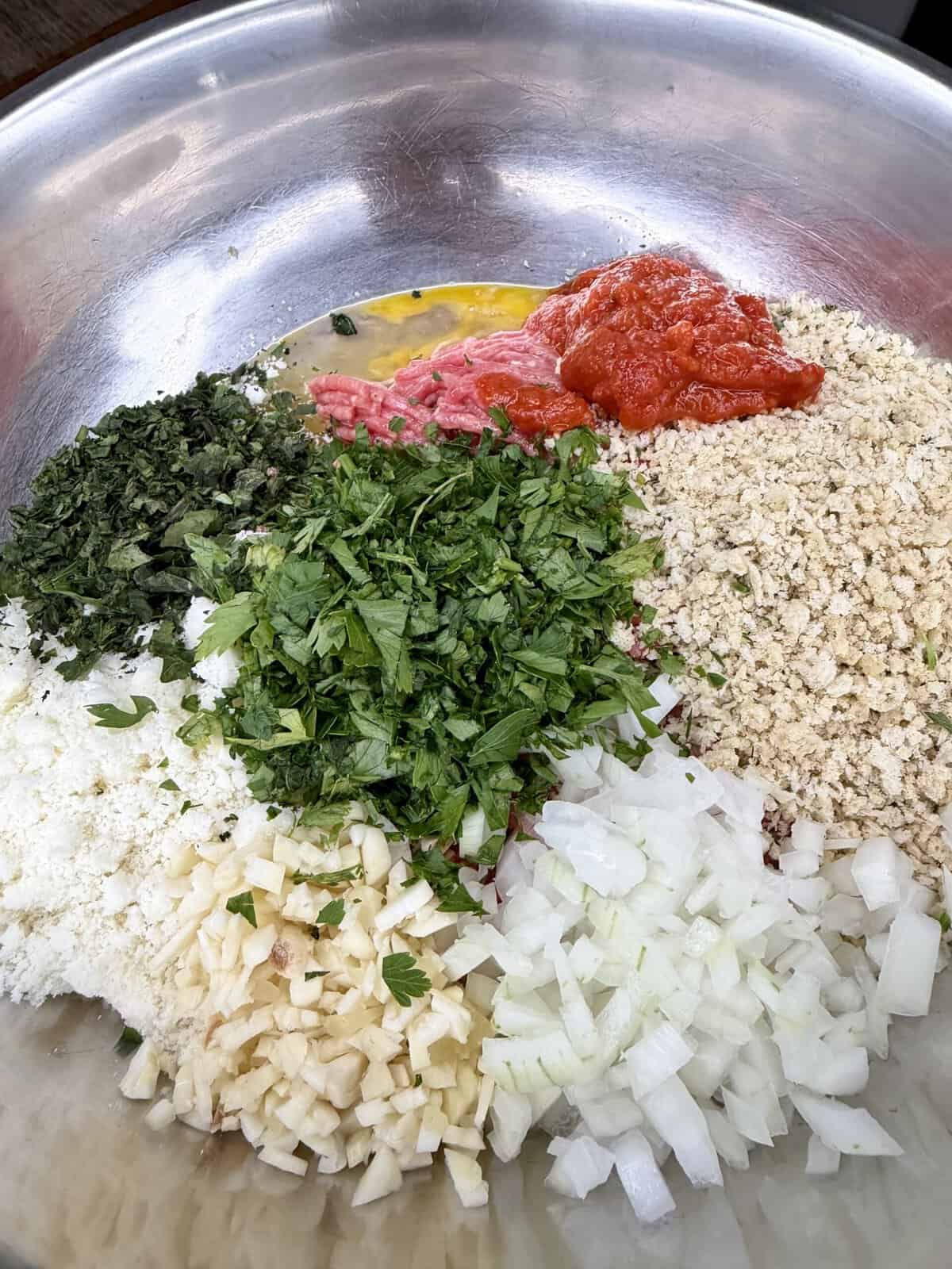 A metal bowl contains chopped parsley, chopped onion, minced garlic, dried herbs, tomato sauce, breadcrumbs, shredded cheese, egg, and ground meat, all arranged in separate piles, ready to be mixed.