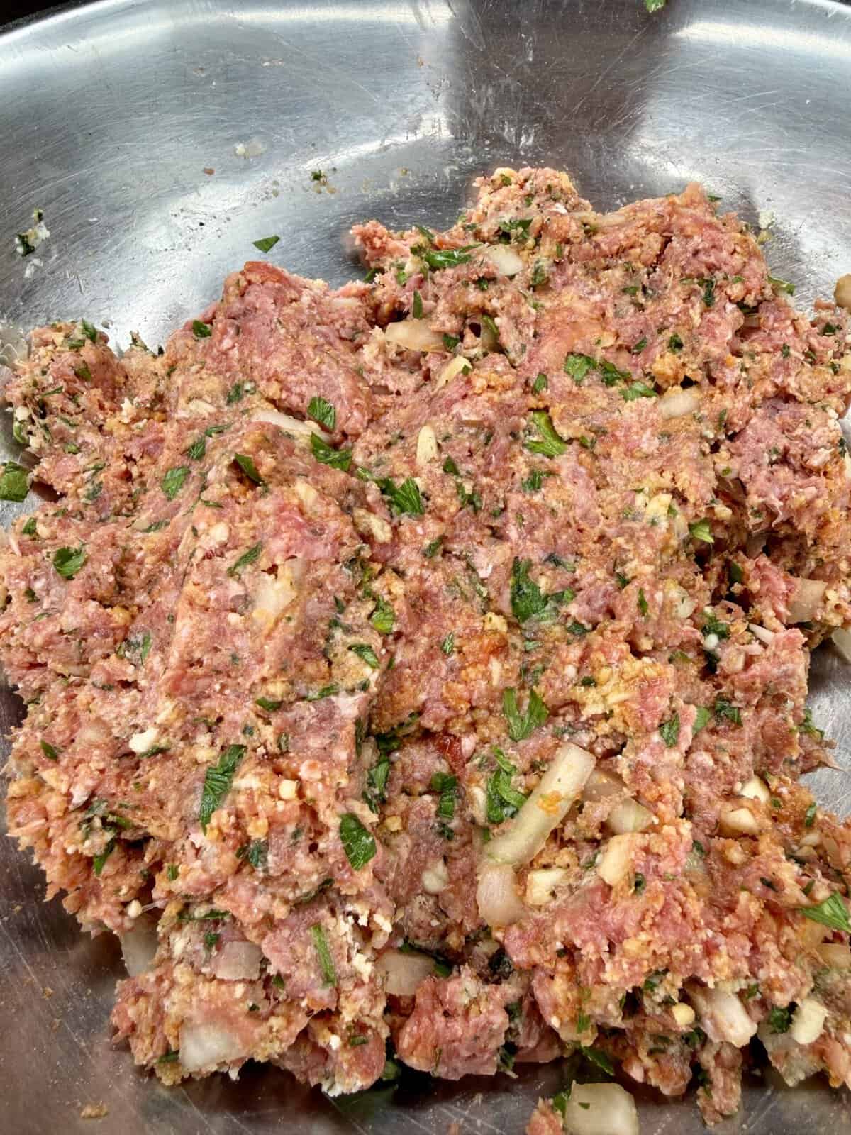 A close-up of a metal bowl filled with raw ground meat mixed with chopped onions, herbs, and spices, ready to be cooked or formed into patties.
