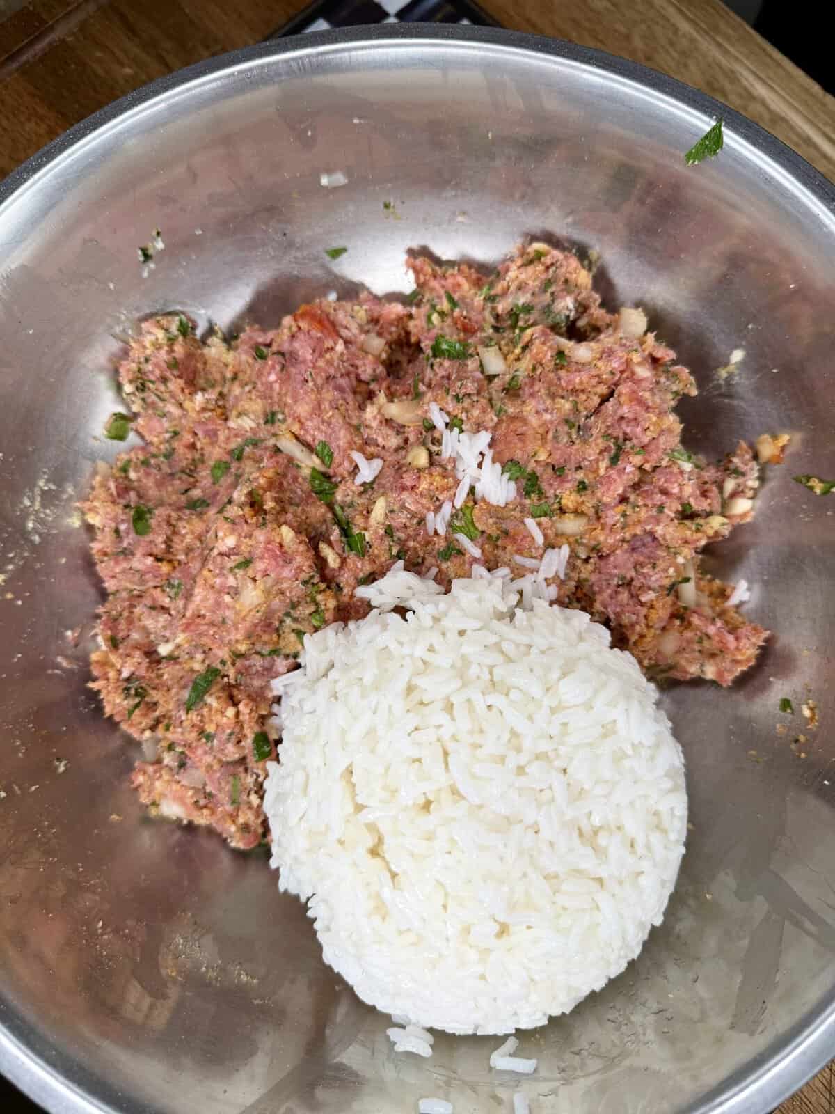 A metal bowl containing a mixture of ground meat, herbs, and spices, with a mound of cooked white rice placed on top of the mixture.
