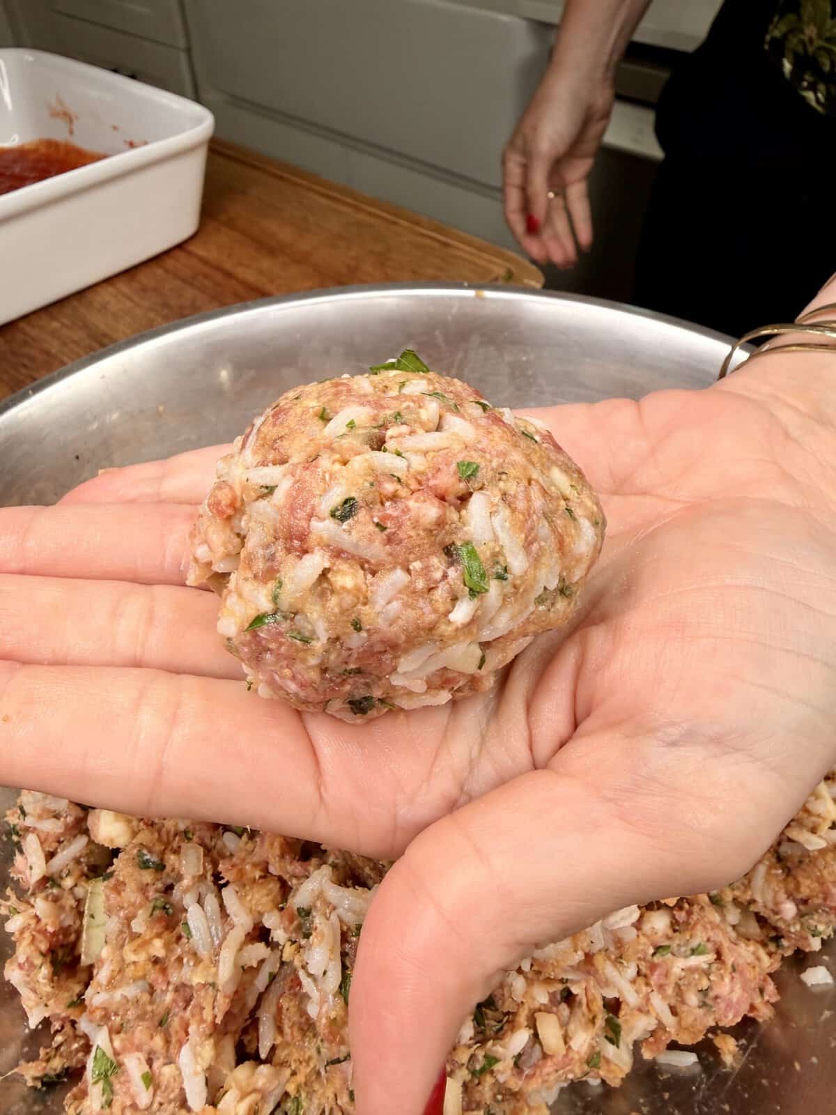A close-up of a hand holding a large, uncooked meatball mixture with rice, herbs, and ground meat over a metal bowl filled with more mixture. Another hand rests on a wooden countertop in the background.