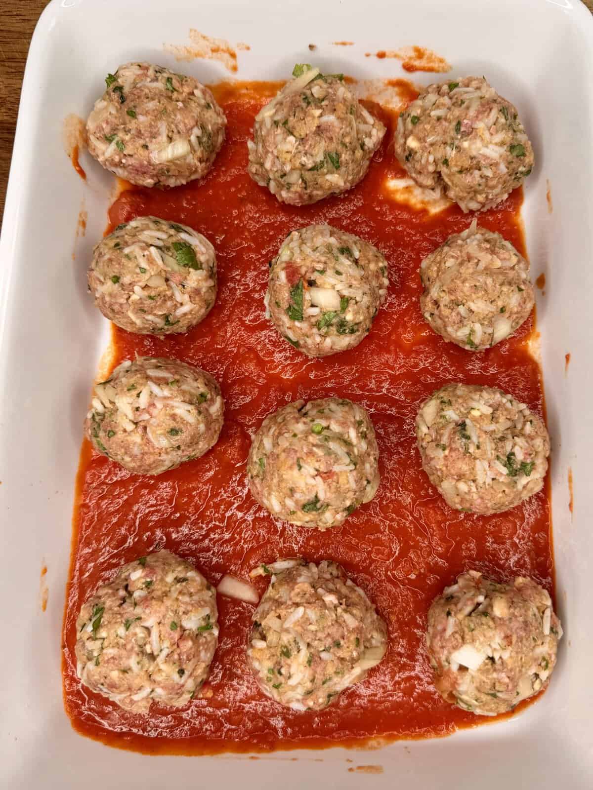 A white baking dish with twelve raw meatballs, mixed with chopped onions and herbs, arranged on a layer of tomato sauce.