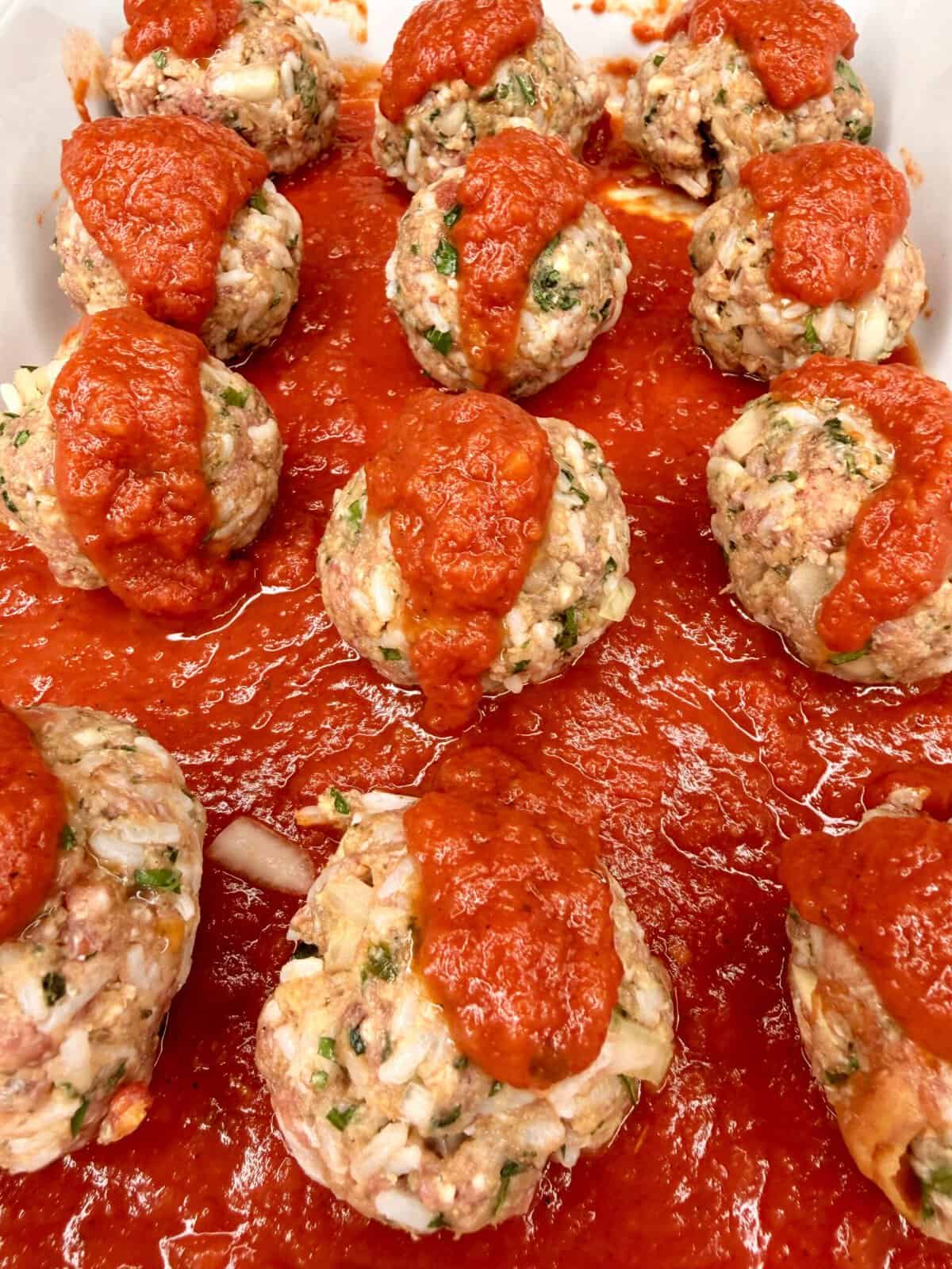 Raw meatballs with chopped onion and herbs are arranged in a baking dish, covered with tomato sauce, and ready to be cooked. Some sauce is spooned on top of each meatball, with more spread across the bottom of the dish.