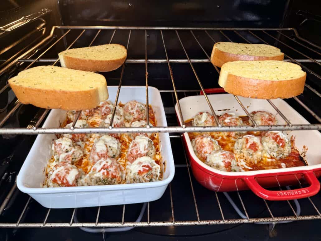Two dishes of cheesy baked meatballs in tomato sauce sit on the middle oven rack, while four slices of bread toast on the top oven rack.