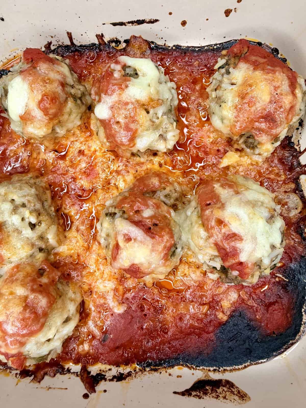 Seven baked meatballs topped with melted cheese and marinara sauce sit in a baking dish. The edges of the sauce are browned and caramelized, giving a slightly crispy appearance around the dish.
