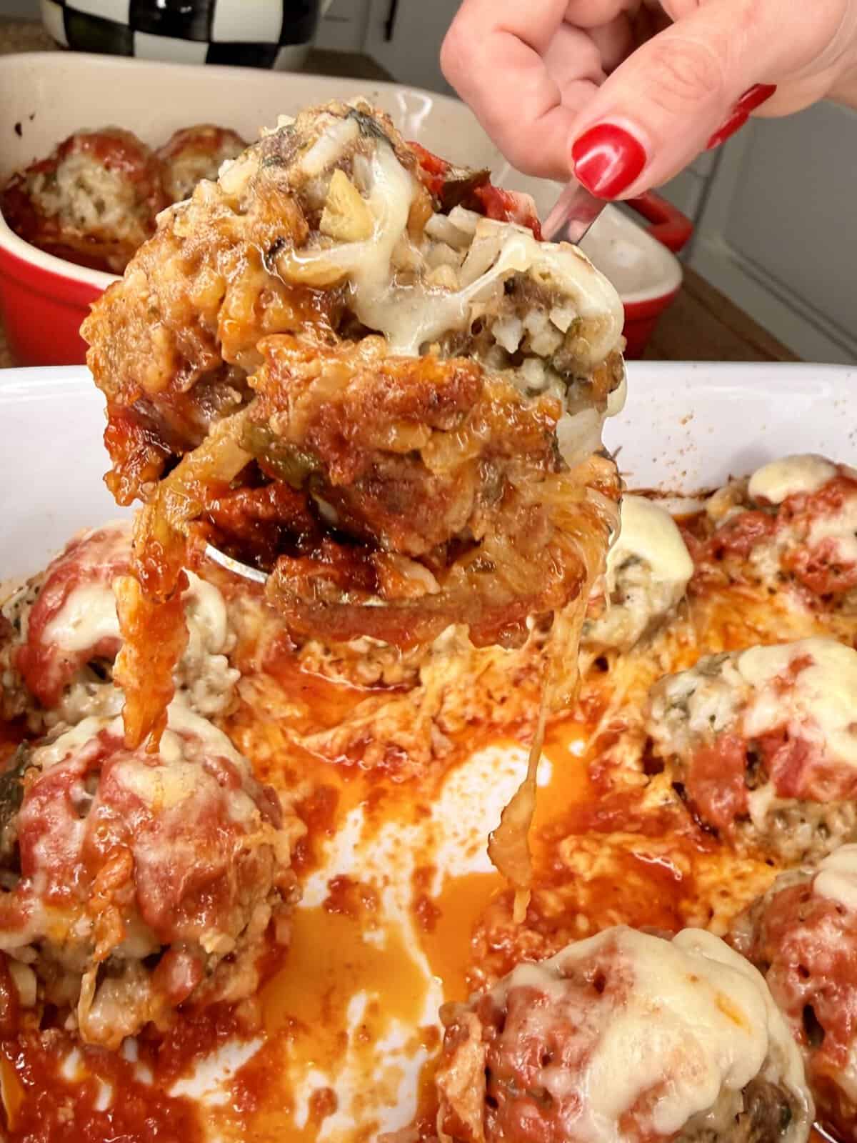 A hand lifts a cheesy, saucy stuffed meatball from a white baking dish filled with similar meatballs topped with melted cheese and tomato sauce.