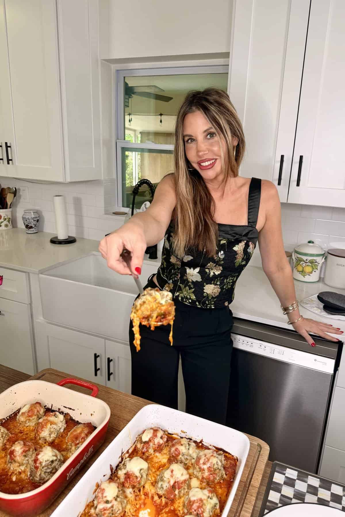 A woman in a black floral top smiles in a kitchen, holding up a serving of cheesy baked meatballs from a casserole dish on a wooden counter. Another dish of meatballs is also visible.