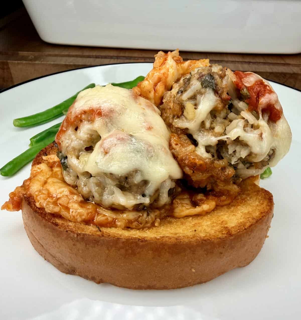 Two cheesy baked meatballs topped with marinara sauce sit on a thick slice of toasted bread, with green beans in the background on a white plate.