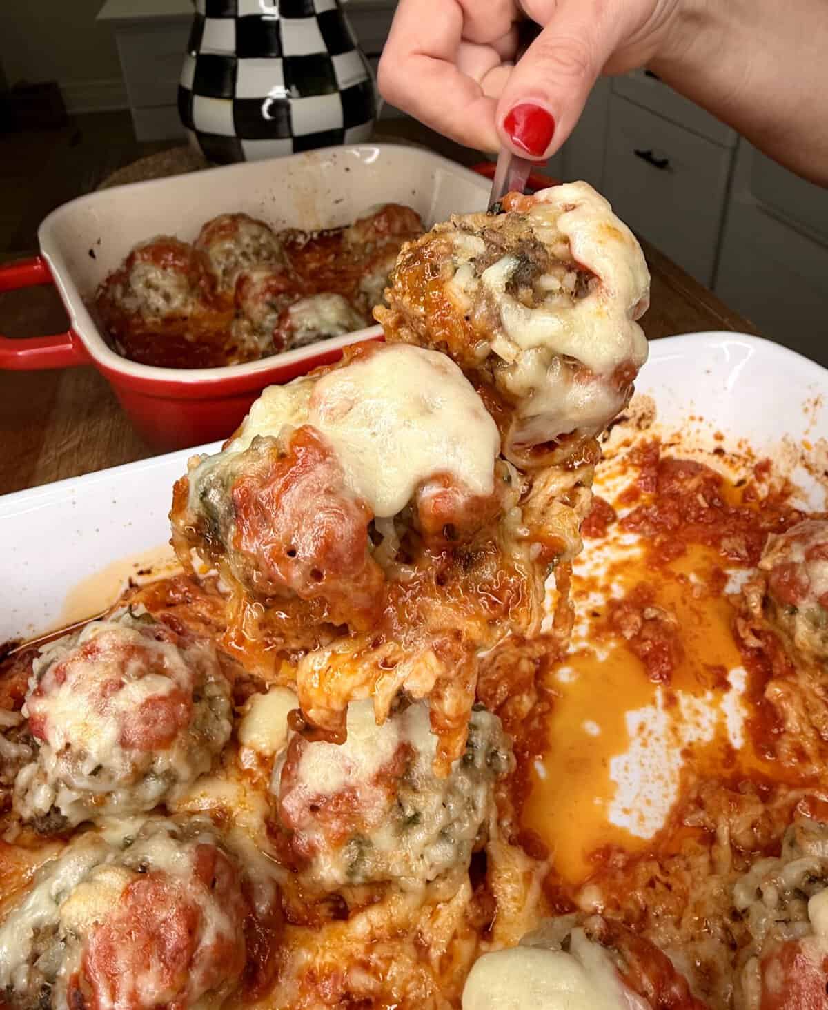 A hand with red nails lifts a cheesy, baked meatball covered in marinara sauce from a casserole dish filled with similar meatballs. A second dish with more meatballs sits in the background.