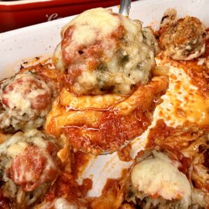 A close-up of a cheesy stuffed pepper being lifted from a baking dish, surrounded by several other stuffed peppers topped with melted cheese and tomato sauce.