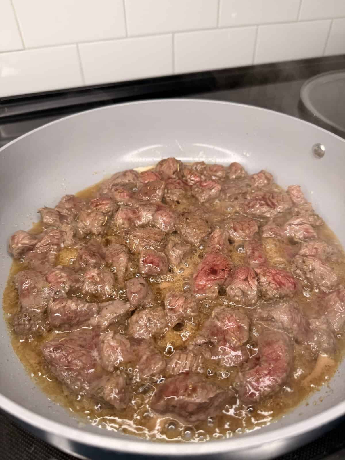Chunks of beef simmer in a frying pan with brown sauce, evoking the comforts of Best One Pot Steak and Rice, as they cook over a stovetop. White subway tile backsplash is visible in the background.
