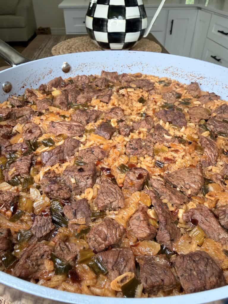 A large skillet filled with Best One Pot Steak and Rice featuring chunks of beef, rice, onions, celery, and green peppers in a savory sauce, with a checkered teapot in the background.