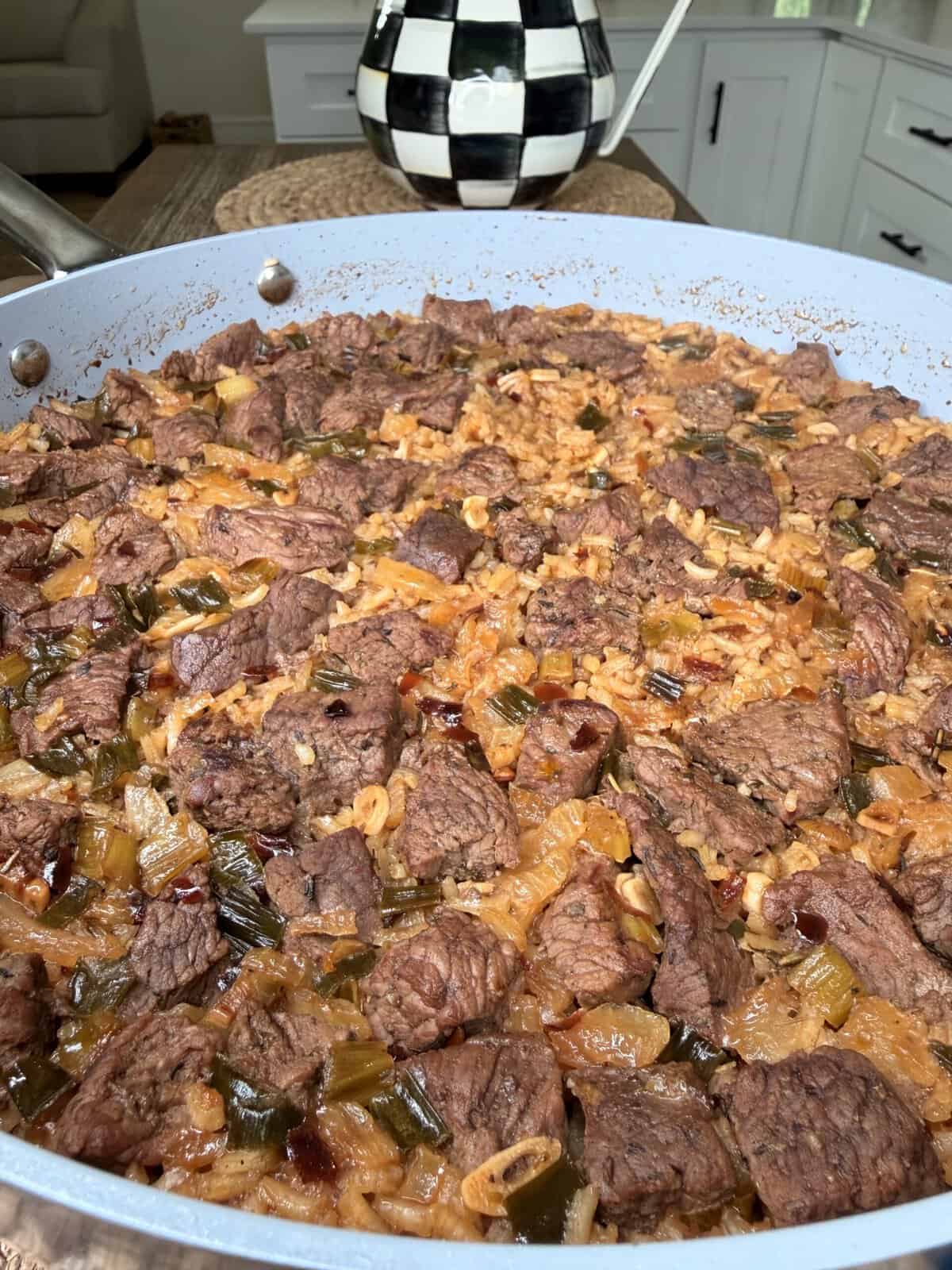 A large skillet filled with Best One Pot Steak and Rice featuring chunks of beef, rice, onions, celery, and green peppers in a savory sauce, with a checkered teapot in the background.