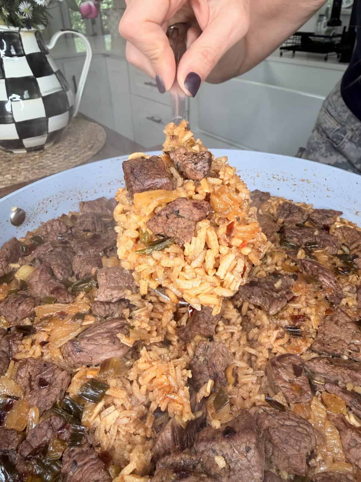 A hand holding a serving spoon scoops up a portion of the Best One Pot Steak and Rice from a pan filled with tender beef chunks, fluffy rice, onions, and seasonings in a cozy kitchen setting.