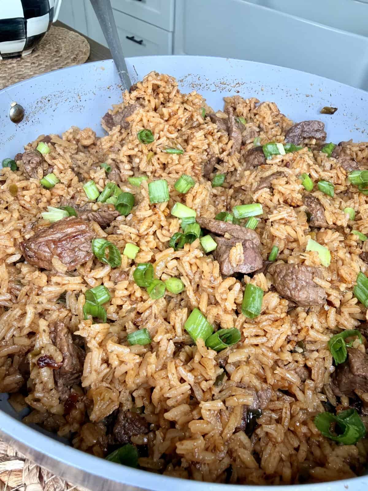 A close-up of a pan filled with cooked rice, chunks of browned beef, and chopped green onions, with a metal utensil resting in the pan. This savory Best One Pot Steak and Rice dish is beautifully garnished with herbs.