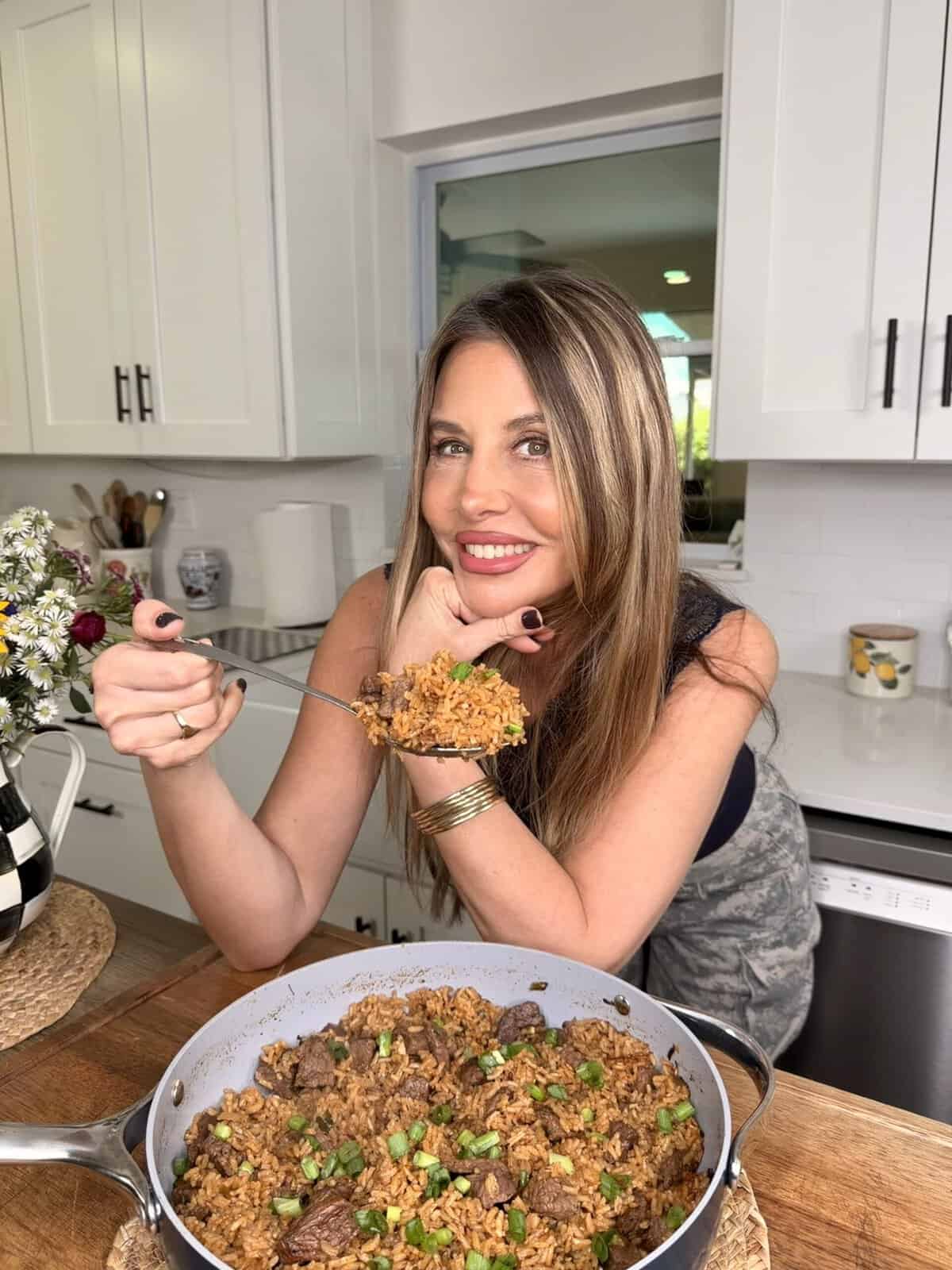 A smiling woman with long hair enjoys a forkful of Best One Pot Steak and Rice from a pan in a modern kitchen with white cabinets and fresh flowers on the counter.
