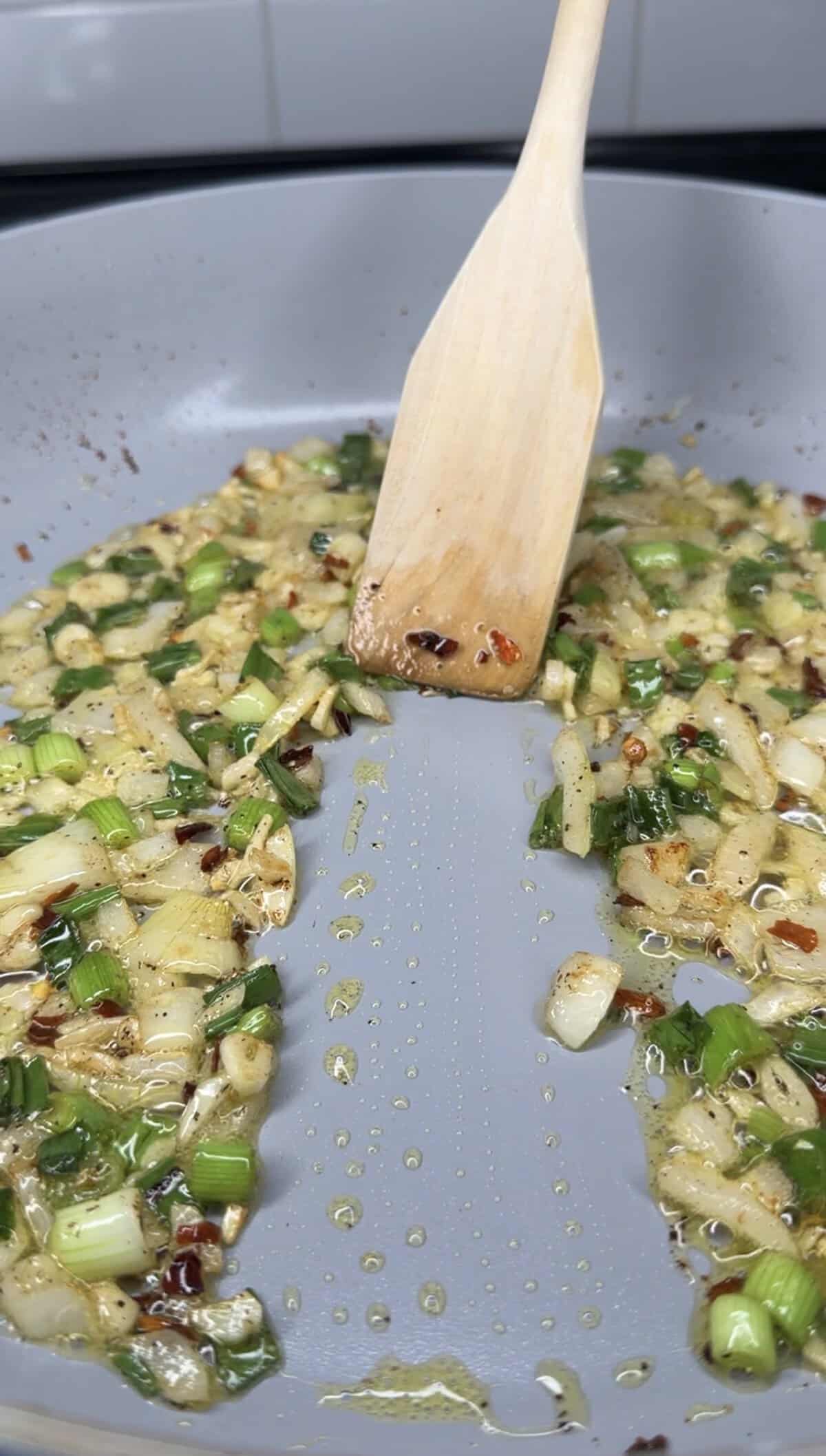 Chopped onions and green onions are being sautéed with red pepper flakes in oil in a pan, forming the flavorful base for the Best One Pot Steak and Rice. A wooden spatula stirs the mixture, leaving a gap in the center.