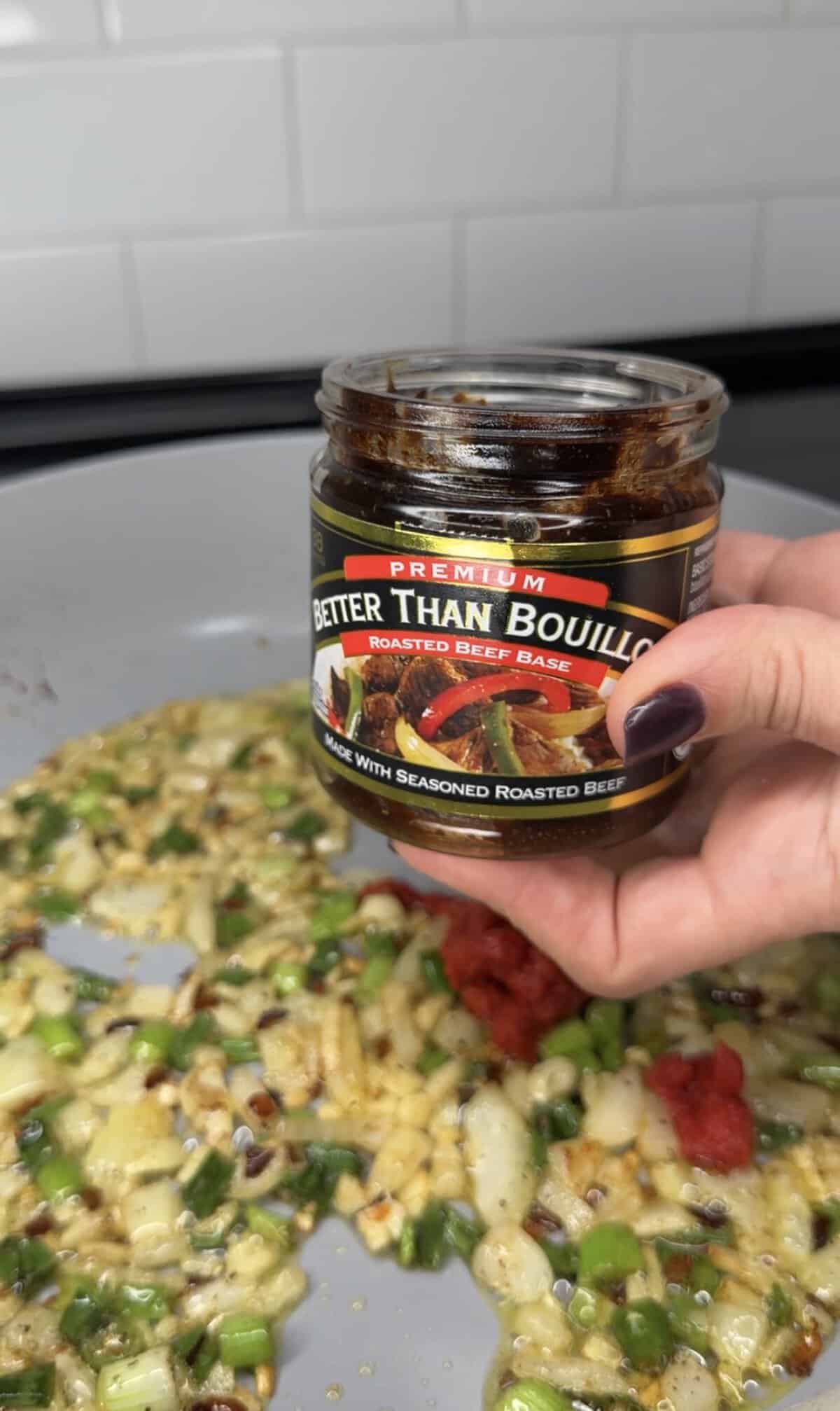 A hand holds a jar of Better Than Bouillon roasted beef base over a pan filled with sautéed onions, green onions, and tomato paste—the perfect start for the Best One Pot Steak and Rice. The kitchen backsplash is visible in the background.