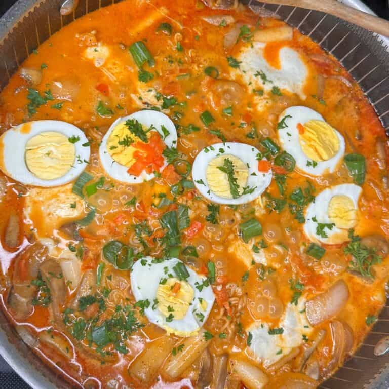 A close-up of a pot of spicy orange soup with sliced boiled eggs on top, garnished with chopped green onions, fresh herbs, and diced red peppers. Vegetables and pasta are visible in the broth.