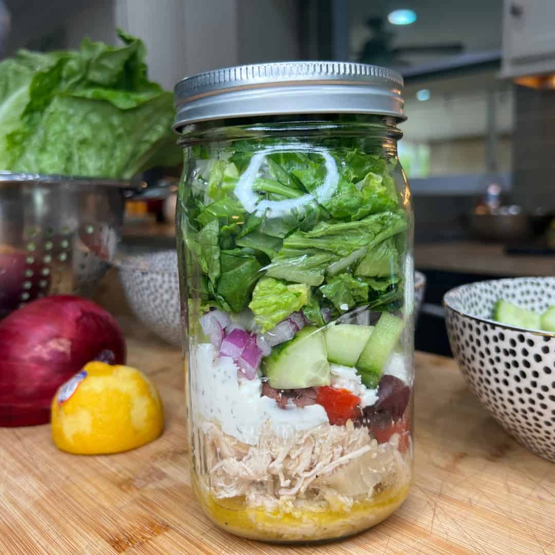 A mason jar filled with layered salad ingredients, including shredded chicken, dressing, cherry tomatoes, olives, cucumbers, red onions, and chopped lettuce. The jar sits on a wooden counter with vegetables in the background.